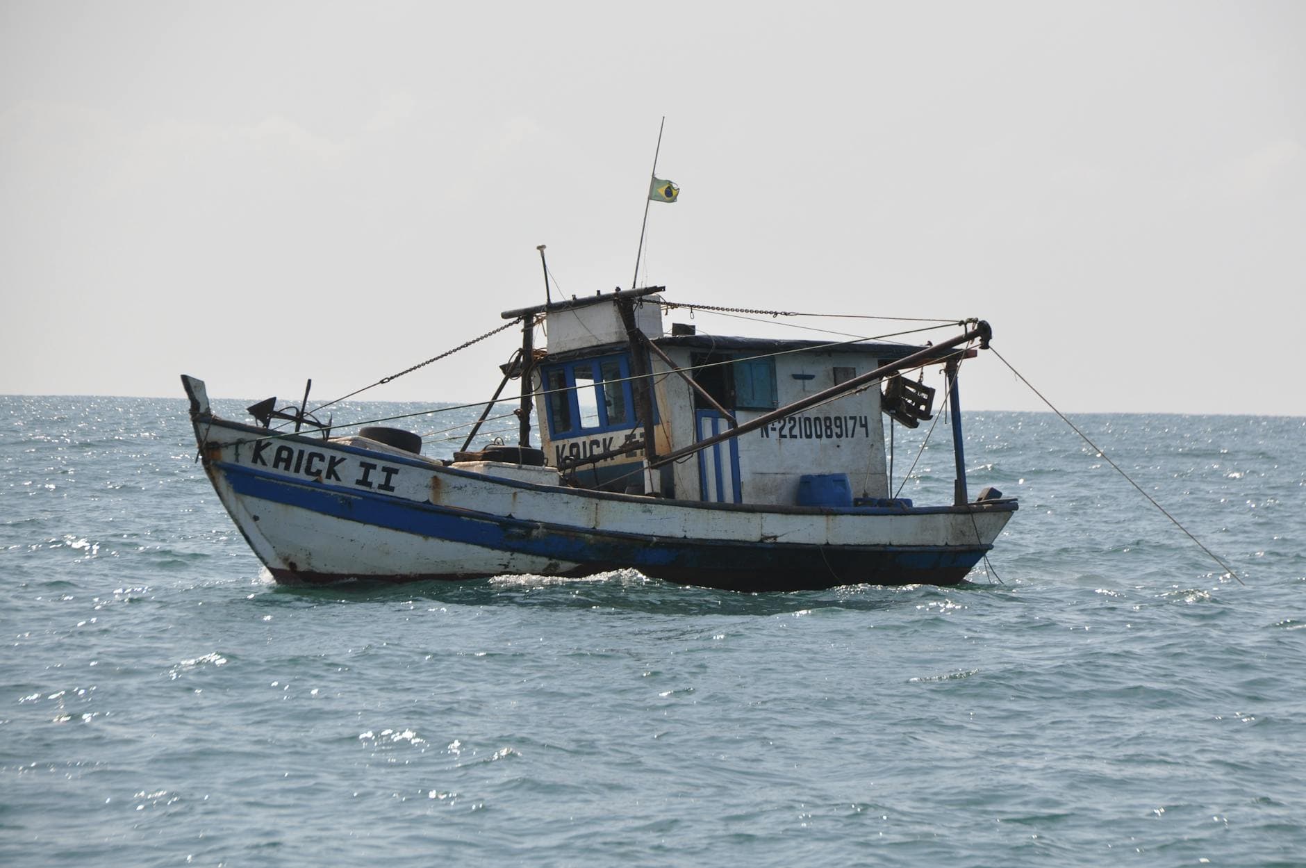 A rustic fishing boat flying the Brazilian flag is captured sailing on the open sea with a clear horizon.