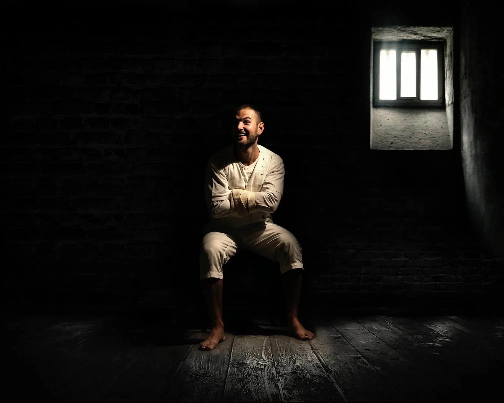A man in a dimly lit room wearing a straightjacket, sitting with a smile in an asylum setting.