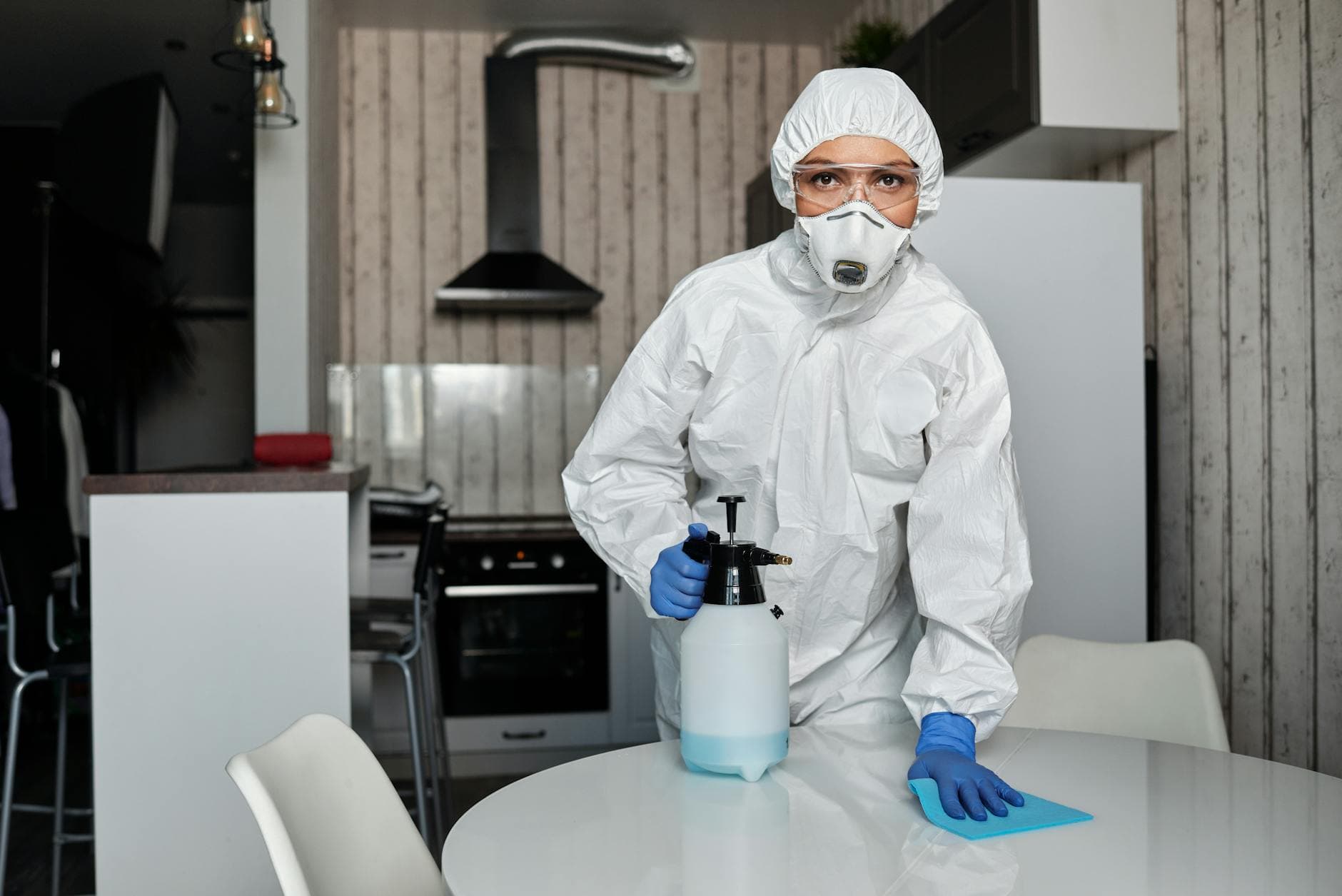 A cleaner in full protective gear disinfects a table indoors, ensuring a sterile environment.