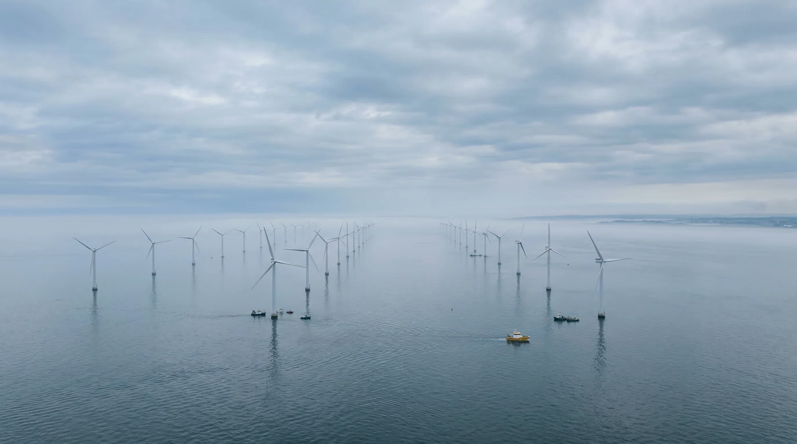 A wide-angle landscape shot of offshore wind turbines standing in the calm, grey-blue waters of the Baltic Sea under a soft, overcast sky. The turbines are arranged in a geometric pattern extending to