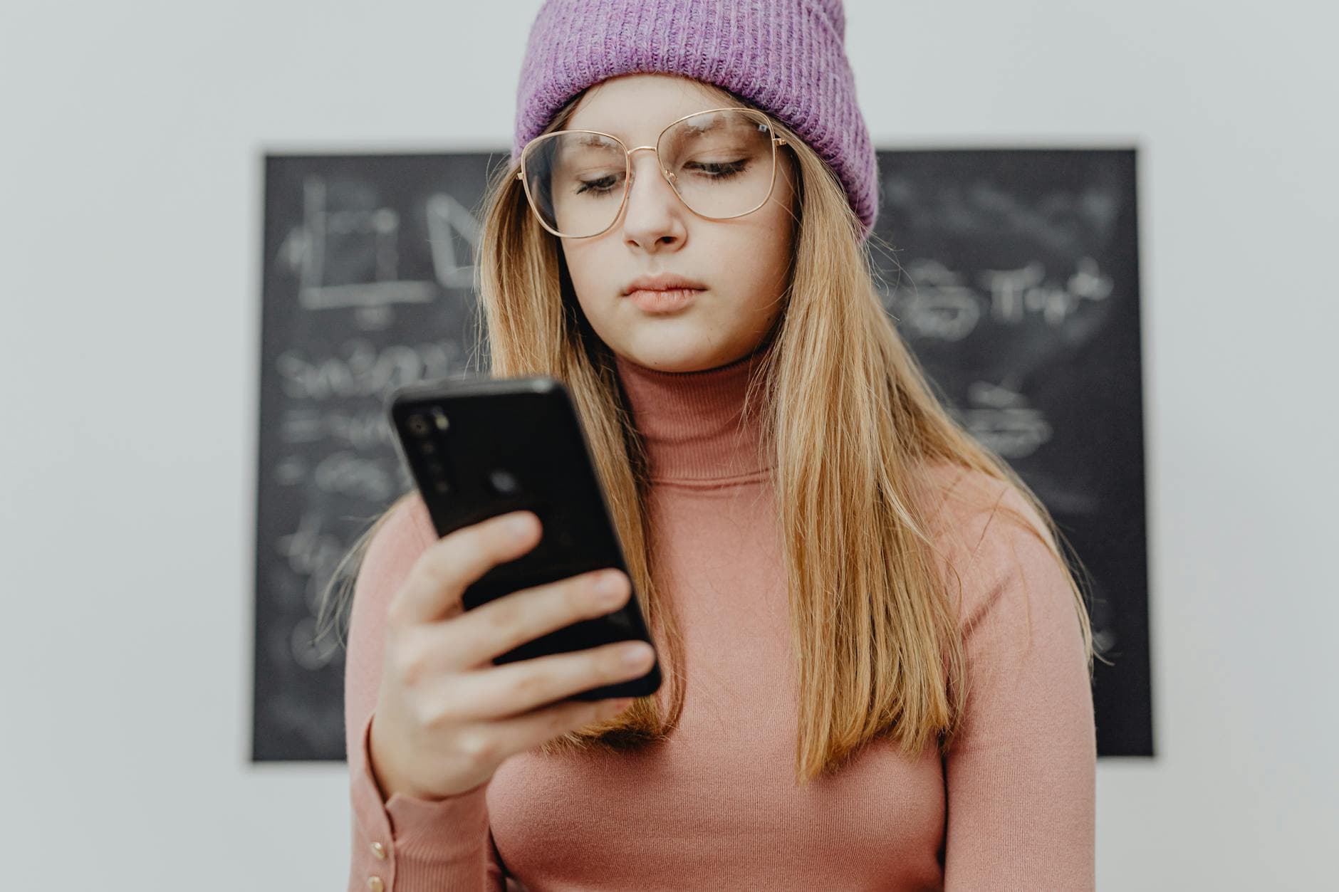 Teenage girl wearing glasses and knit hat using a smartphone indoors.