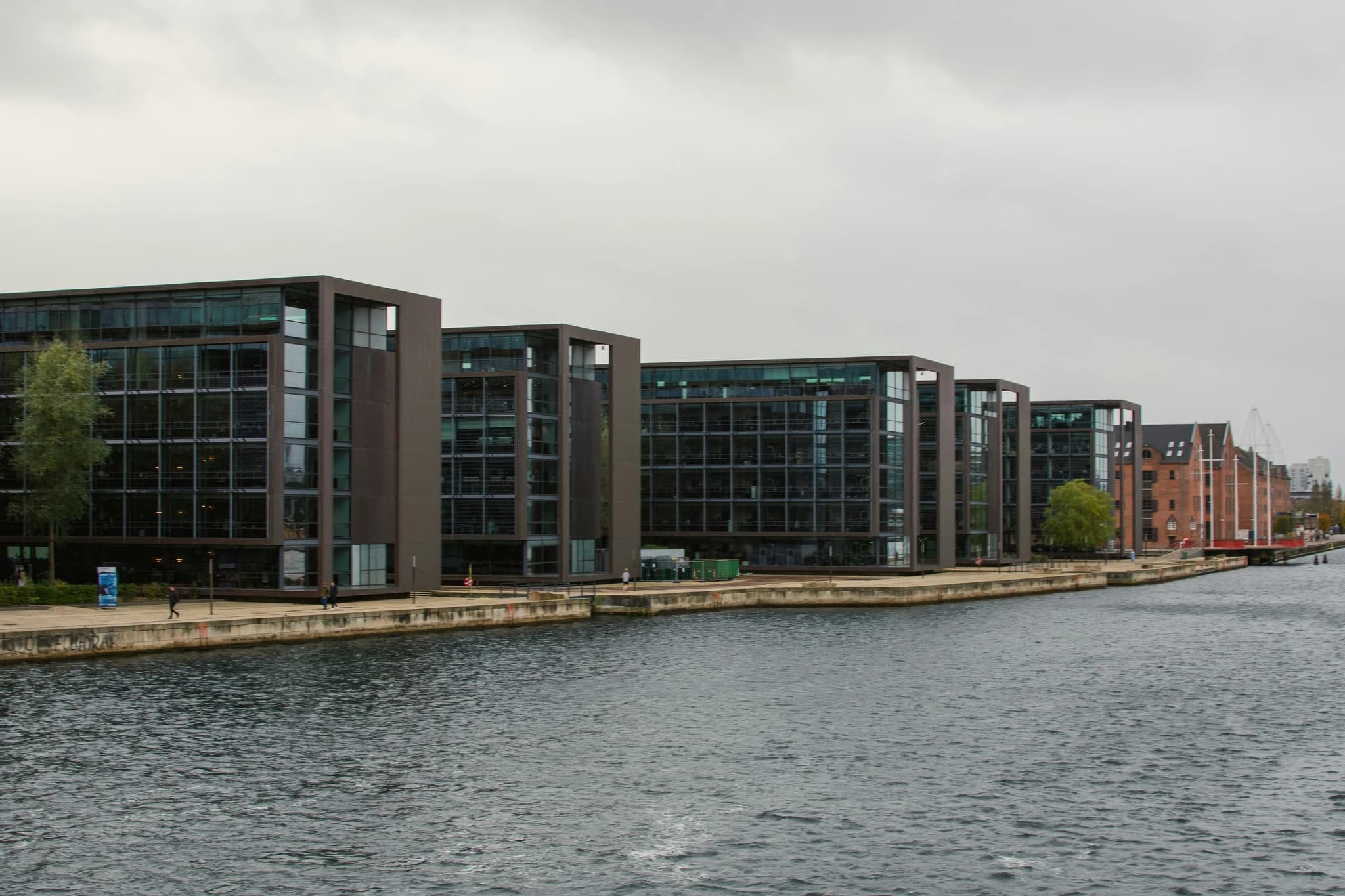 Modern buildings line a calm canal under a cloudy sky.
