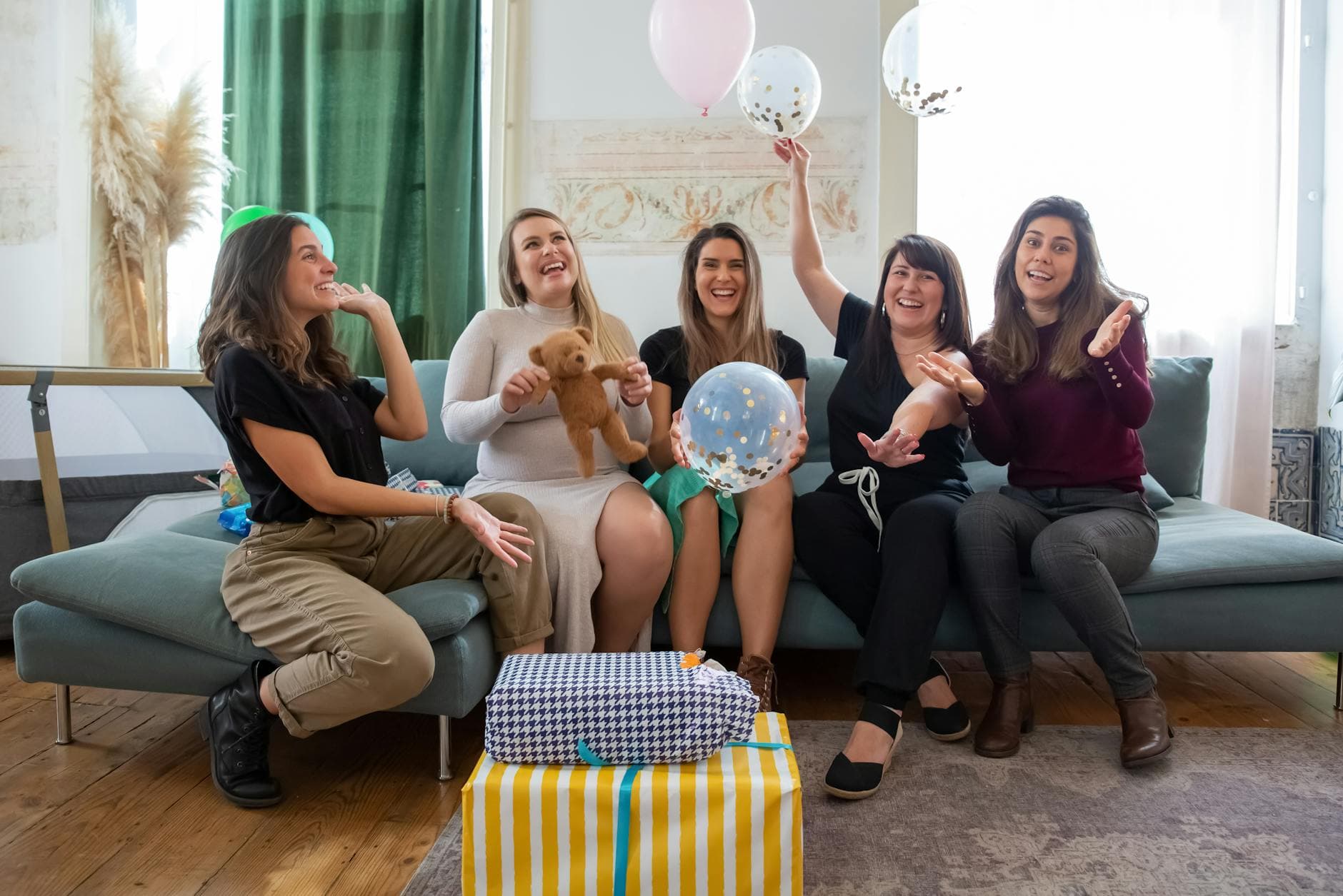 Five friends enjoy a joyful baby shower with gifts and balloons indoors, celebrating together.